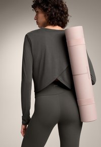 Henkilö tummanharmaassa urheiluvaatteessa kantaa rullattua vaaleanpunaista joogamattoa olkapäillään, nähtynä takaa.