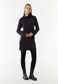 Strickkleid in Dunkellila mit einem Schalkragen, geripptem Saum und strukturierten Ärmeldetails, kombiniert mit schwarzen Strumpfhosen und klobigen schwarzen Stiefeln.