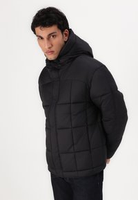 Zwarte pufferjas met capuchon, voorzien van een geruite ontwerp en een voorrits. Zachte textuur en een iets chunky silhouet.