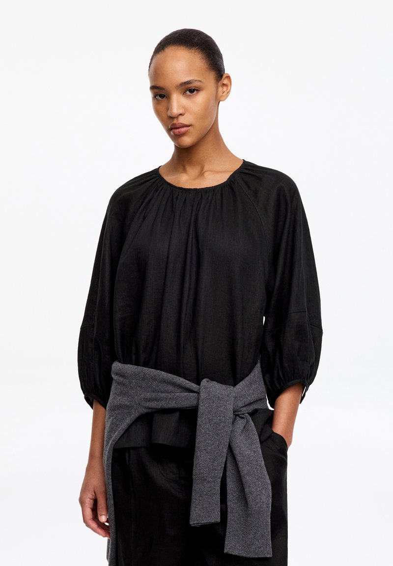 Femme les cheveux attachés, portant un haut noir ample à manches trois-quarts et un pull gris noué autour de la taille.