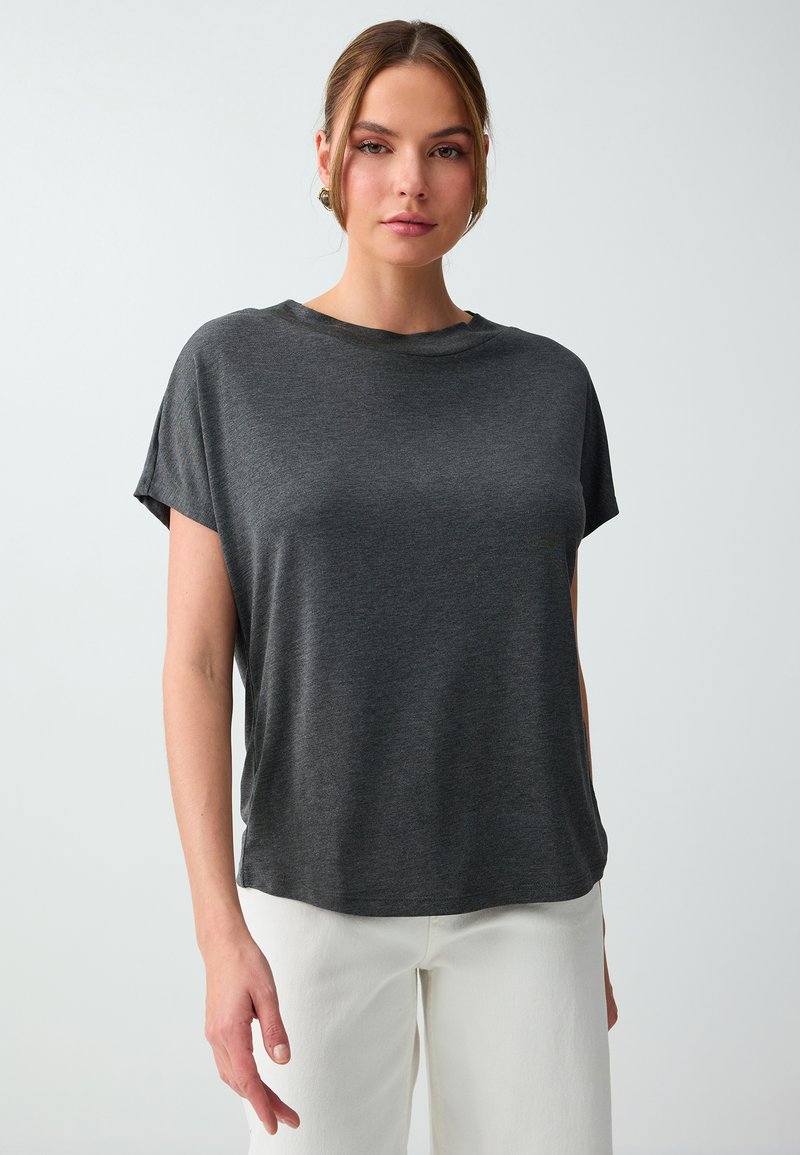 Jeune femme aux cheveux châtain clair portant un t-shirt gris foncé à manches courtes et un pantalon blanc, debout devant un fond clair uni.