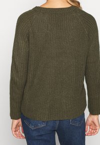 Strickpullover in dunkelolivgrün mit einem strukturierten, gerippten Design. Er hat lange Ärmel und einen runden Halsausschnitt und wird mit blauen Jeans kombiniert.