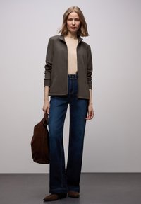 Chaqueta de cremallera de color verde oscuro de corte relaxed sobre una blusa beige, combinada con vaqueros azules de cintura alta; llevando un bolso tote de ante marrón.