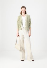  Lichtgroene gebreide cardigan met knopen, gecombineerd met een witte top en wijde beige broek. Het model houdt een kleine beige handtas vast.