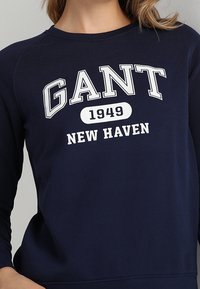 Felpa blu navy con girocollo e maniche lunghe a raglan. Presenta un testo stampato bianco: "GANT 1949 NEW HAVEN" ben visibile.