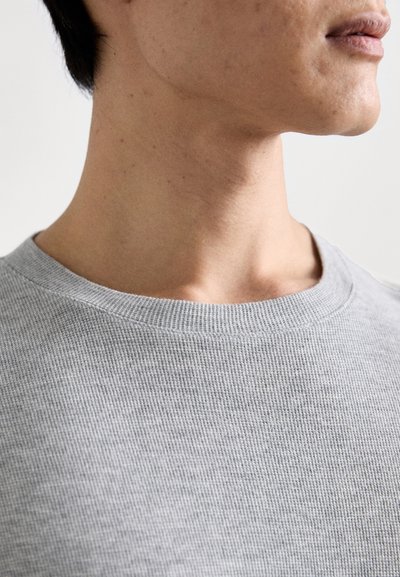 Graues geripptes T-Shirt mit Rundhalsausschnitt, das einen strukturierten Stoff mit weichem Gefühl zeigt. Der Ausschnitt ist weit und sitzt direkt unter dem Schlüsselbein.