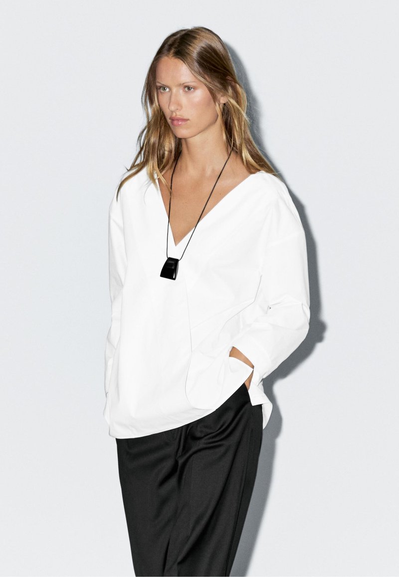 Hvid V-hals bluse lavet af bomuld med lange ærmer, parret med sorte bukser. En sort pendantsmykke tilføjer detalje.