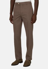 Homme portant un pantalon droit marron et des mocassins en daim marron, debout devant un fond blanc uni.