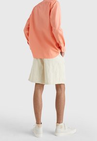 Camicia leggera a maniche lunghe corallo con colletto button-down, abbinata a pantaloni beige. Sneakers bianche alte completano il look.