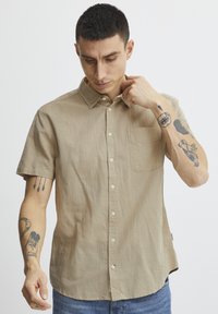 Camisa de botón de manga corta en tono beige, hecha de un tejido ligero, que cuenta con un bolsillo en el pecho y un cuello clásico, con un corte relajado y textura mínima.