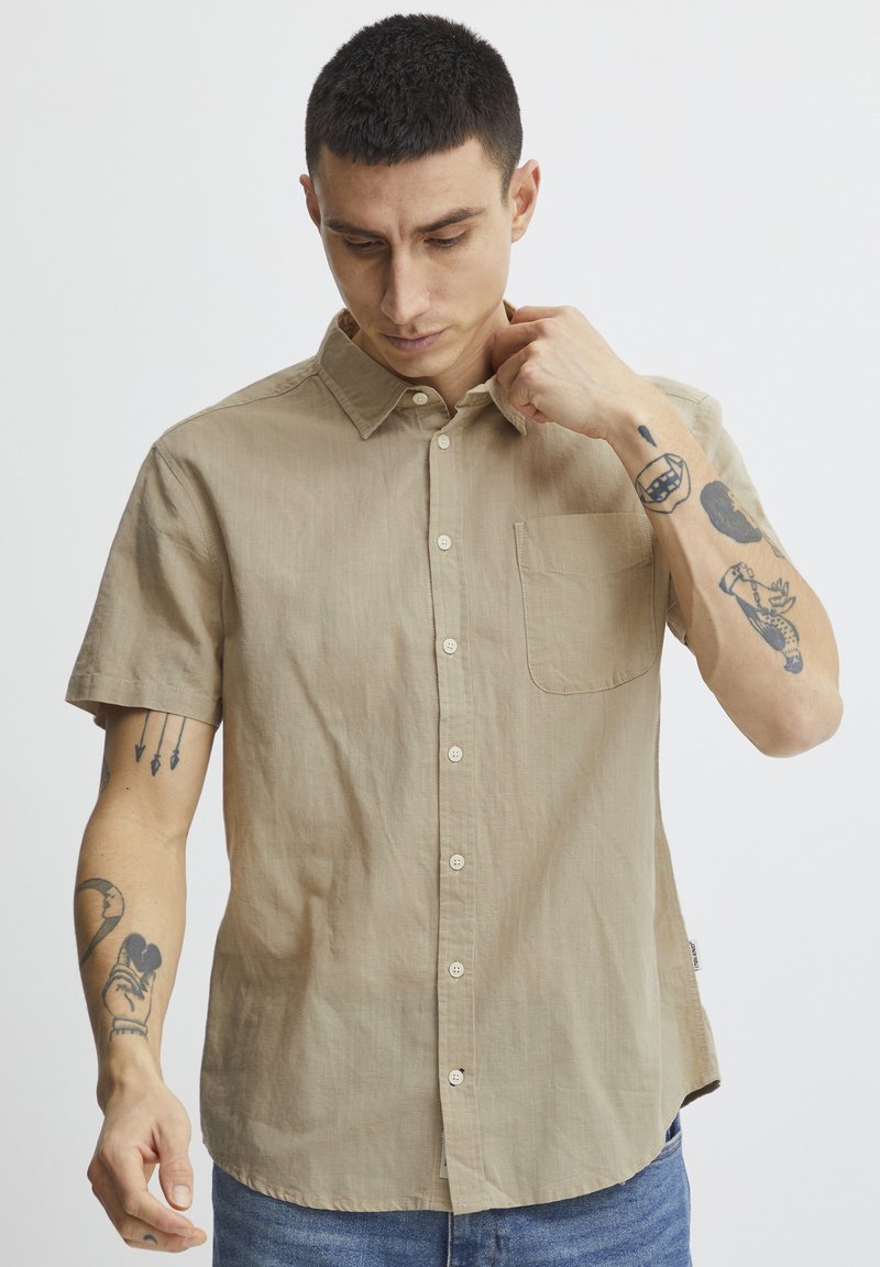 Camisa de botón de manga corta en tono beige, hecha de un tejido ligero, que cuenta con un bolsillo en el pecho y un cuello clásico, con un corte relajado y textura mínima.