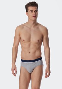 Graue Baumwollunterhosen mit einem marineblauen Bund, der weiße Akzente und Markenlogo aufweist. Das Modell steht mit neutralem Gesichtsausdruck und natürlicher Haltung.