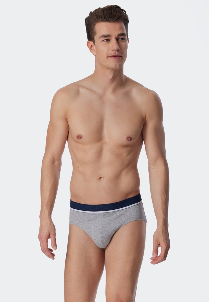 Graue Baumwollunterhosen mit einem marineblauen Bund, der weiße Akzente und Markenlogo aufweist. Das Modell steht mit neutralem Gesichtsausdruck und natürlicher Haltung.