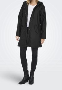 Imperméable noir avec capuche, fermeture à boutons-pression, poches latérales et manches longues. Porté avec un jean noir ajusté et des bottines.