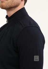 Hombre vistiendo un suéter azul marino con cremallera y un cuello alto, con un pequeño parche rectangular de logo en la parte superior de la manga.