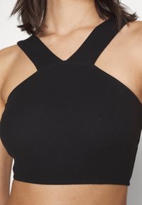 Crop top noir côtelé avec des bretelles épaisses croisées sur les épaules nues et le haut de la poitrine, porté par une personne aux cheveux foncés mi-longs.
