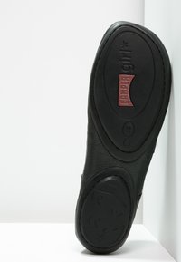 Zapato de cuero negro con suela texturizada que presenta un logo rojo de "Camper" y talla 38. Tacón curvado y bordes suaves para mayor comodidad.