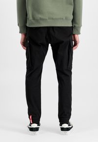 Černé cargo kalhoty s bočními kapsami, zúženými nohavicemi a elastickými manžetami, doplněné černobílými teniskami. Model má na sobě zelenou mikinu.