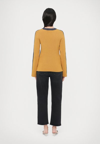 Camisola amarela com nervuras e detalhes em azul-marinho, corte ajustado e mangas compridas, combinada com calças escuras de perna reta e saltos claros.