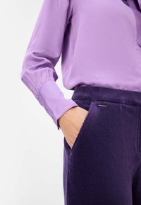 Hedvábná, levandulová košile s vysokým límcem a elastickými manžetami, v kombinaci s tmavě fialovými kordovým kalhotami s bočními kapsami a kovovým akcentem.