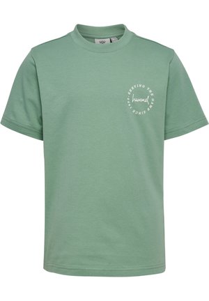 Barn med langt krøllet hår står og smiler, iført en grøn t-shirt og matchende shorts, hvide sokker og sneakers mod en ensfarvet baggrund.