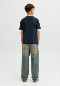 Un garçon aux cheveux courts portant un t-shirt bleu marine, un jean faded ample et des chaussures beiges, se tenant debout, tourné le dos devant un fond uni.