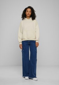 Beige hettegenser med frontlomme, kombinert med vide blå cargo-bukser med sidelommer og hvite sneakers. En enkel, uformell antrekk.