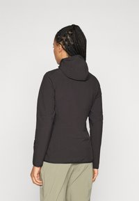 Woman with braided hair wearing a black hooded jacket and light green pants, standing with back facing the camera.