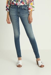 Woman wearing faded blue skinny jeans, a colorful floral blouse, and open-toe white sandals with buckle detail.