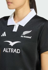 Svart sportströja med vit krage. Har texten: "ALL BLACKS," "ALTRAD," samt en logotyp. Tillverkad av texturerat material, med en åtsittande design.