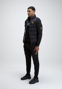 Czarna kamizelka puchowa z logo, z przodu na suwak i kieszeniami bocznymi, w połączeniu z dopasowanymi czarnymi spodniami z kieszeniami cargo i akcentami z logo oraz czarnymi sneakersami.