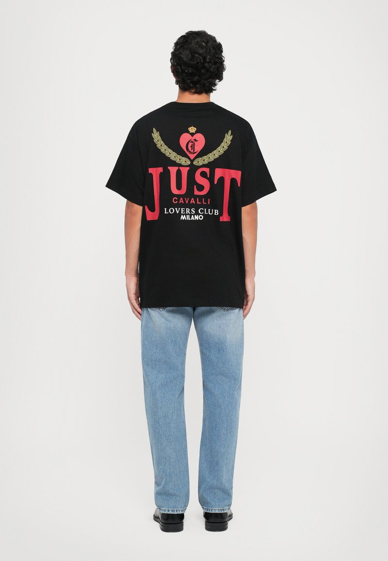 T-shirt de algodão preto com um grande design gráfico nas costas, com letras vermelhas e detalhes em dourado. Combinado com jeans azul claros e sapatos pretos.