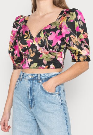 Vrouw draagt een zwart cropped bloesje met bloemenprint en pofmouwen, gecombineerd met lichtblauwe, hooggetailleerde jeans. Ze heeft één hand in haar zak, tegen een egale achtergrond.