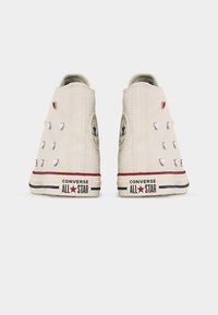 Λευκά sneakers Converse υψηλής κορυφής με κόκκινες και ασημένιες καρδιόσχημες τρουκς, εμφανιζόμενα από πίσω σε λευκό φόντο.