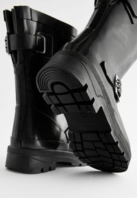 Bottes en caoutchouc noires avec une semelle épaisse et texturée, accents de boucle sur le côté et une finition lisse. Le design présente une hauteur au milieu du mollet et des bords arrondis.