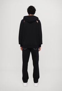 Haut à capuche noir avec cordon de serrage et coupe oversize, associé à un pantalon noir avec des accents cousus. Le fond est uni et clair.