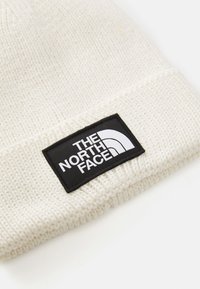 Baltas megztas kepurė su sulenkta rankove, kuri turi juodą stačiakampį lopą su balta tekstine ir grafine The North Face logo.