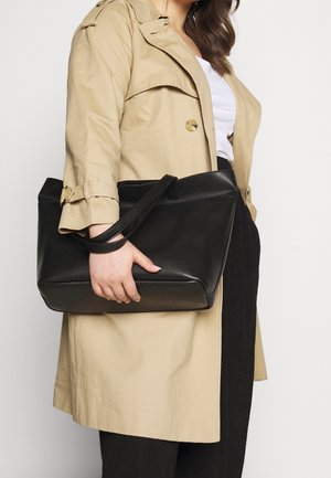 Personne portant un trench-coat beige et un pantalon noir tenant un grand sac fourre-tout en cuir noir avec des bretelles d'épaule.
