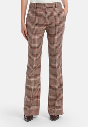 Vrouw in hoge taille bruine en rode geruite wijde broek met zakken, gecombineerd met donkere puntige schoenen, staand tegen een witte achtergrond.