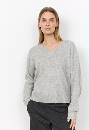Frau mit langen blonden Haaren, die einen hellgrauen Pullover mit V-Ausschnitt und dunkelgraue strukturierte Hose trägt und mit einer Hand in der Tasche steht.
