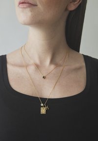 Geschichtete goldene Halsketten mit verschiedenen Anhängern: einer rechteckigen mit einem wiegenden Design, einem sternförmigen und einem Buchstaben "M".