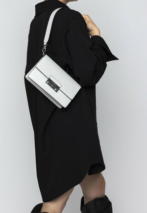 Borsa in pelle bianca di forma rettangolare, con hardware argentato, una patta frontale e tracolla regolabile, indossata sopra un indumento nero.