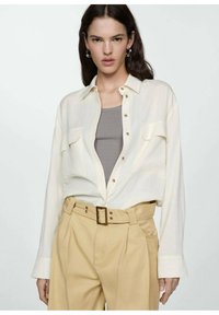 Crème button-up shirt met twee borstzakken, gedragen over een grijze tanktop. Gecombineerd met lichtbeige high-waisted broeken en een riem.