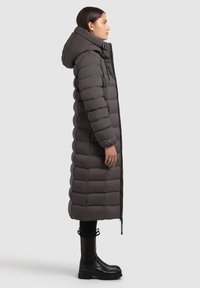 Cappotto imbottito grigio con un design lungo e isolato, dotato di cappuccio, motivo a righe verticali e cerniera frontale. Stivali neri al ginocchio visibili.