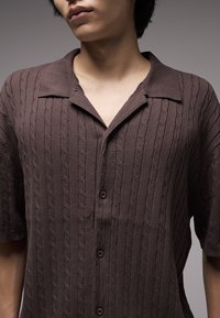 Καφέ κοντομάνικο πουκάμισο με πλεκτό σχέδιο καλώδιου, κουμπιά μπροστά και γιακά. Μαλακή υφή και άνετη εφαρμογή.