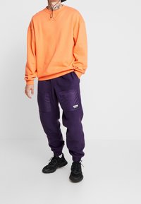Pomarańczowa bluza z długimi rękawami i okrągłym dekoltem, zestawiona z fioletowymi dresami z polaru z dużymi bocznymi kieszeniami. Całość dopełniają czarne trampki.