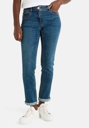 Jean en denim bleu foncé avec un design ajusté, revers roulés et détails de couture subtils, associés à des baskets blanches.
