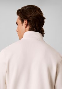 Homme aux cheveux bruns vu de dos, portant une chemise blanche à col montant sur un fond gris clair uni.