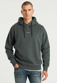 Felpa con cappuccio verde scuro realizzata in tessuto morbido, con tasca frontale e cappuccio regolabile con cordino. Piccolo logo stampato sul petto. Jeans indossati sotto.