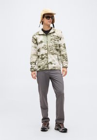 Νεαρό άτομο που φοράει μπουφάν φλις καμουφλάζ, γκρι παντελόνι, παπούτσια πεζοπορίας, γυαλιά ηλίου και μπεζ χειμερινό καπέλο με καλύμματα αυτιών, στέκεται μπροστά σε ουδέτερο φόντο.